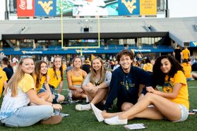 A group of students sitting on a football field wearing blue and gold WVU shirts