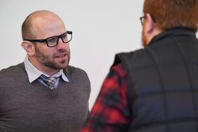 Man in grey sweater and black thick glasses on left. Man in a black vest, red and black plaid shirt on the right