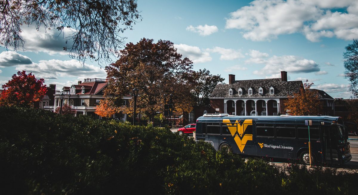 A blue charter bus with a gold Flying WV sits on the street in front of the Mountainlair. In the background is E. Moore Hall.