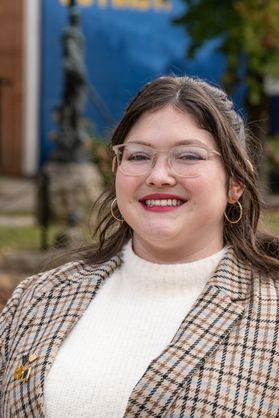 Headshot of WVU student Rachel Johnson. She is pictured outside wearing a brown plaid jacket over a white, mock-neck sweater. She has long brown hair pulled back and wears glasses. 
