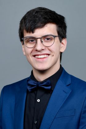Headshot of WVU Bucklew Scholar Brent Marcus. He is pictured against a gray background and is wearing a royal blue jacket with a black dress shirt and royal blue bowtie. He has short black hair and is wearing black-framed glasses. 