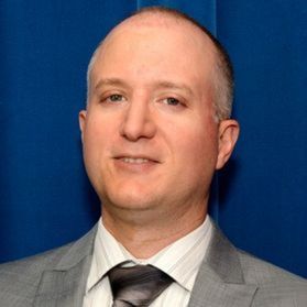 Headshot of WVU researcher Ed Timmons. He is pictured in front of a blue background wearing a gray suit over a white dress shirt and dark colored tie. He has a receding hairline with a bit of gray hair on the sides. 