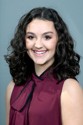 Headshot of WVU Bucklew Scholar Isabella Bottini. She is pictured against a gray background and is wearing a maroon and black blouse with a bow tied at the neck. She has long, curly black hair. 