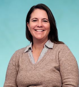 Headshot of WVU professor Lesley Cottrell. She is standing in front of a cream colored wall and is wearing a navy blue cardigan over a white dress shirt. She has shoulder length brown hair. 