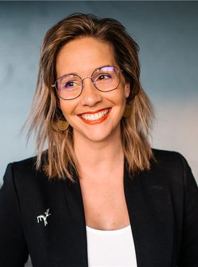 Headshot of Ascend participant Heather Mae Pusztai. She is pictured in front of a blue marbled background wearing a black jacket over a white shirt. She has shoulder length blond hair and wears glasses. 
