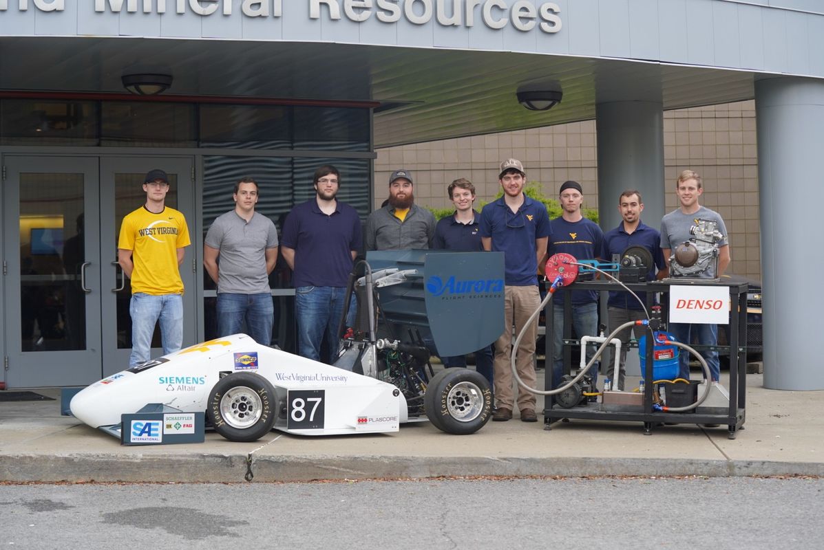 Nine men stand in front of a building showcasing the race car that they built