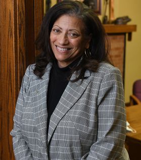 A woman with long dark hair stands in a doorway while wearing a dark shirt under a gray plaid blazer