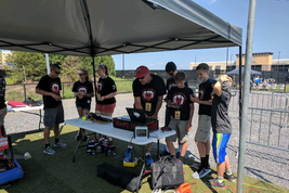 Earl Scime and his young Mountaineer Area Robotics team work outside under a tent. They are standing at a table with computer equipment on top. There are eight individuals in the photograph.