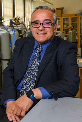 Headshot of WVU researcher Luis Arroyo. He is pictured in the lab wearing a dark jacket over a royal blue dress shirt with a blue and white floral tie. He has short gray hair and wears square, black-framed glasses. 