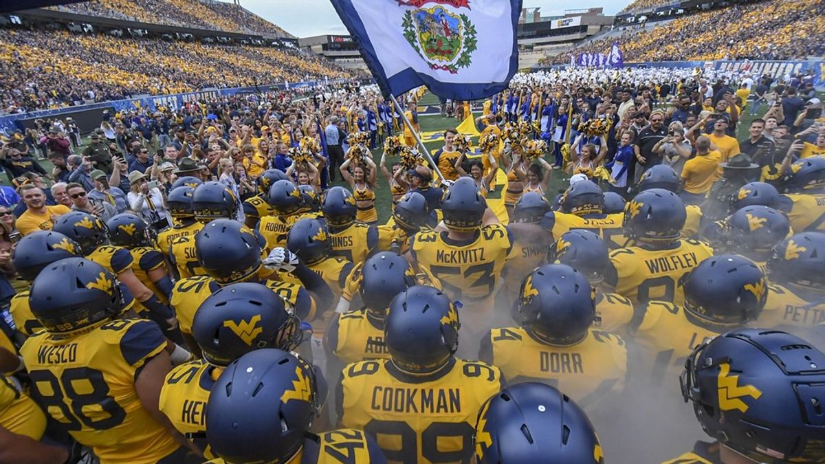 WVU football players stand at Milan Puskar stadium holding the state flag in yellow jerseys with blue helmets
