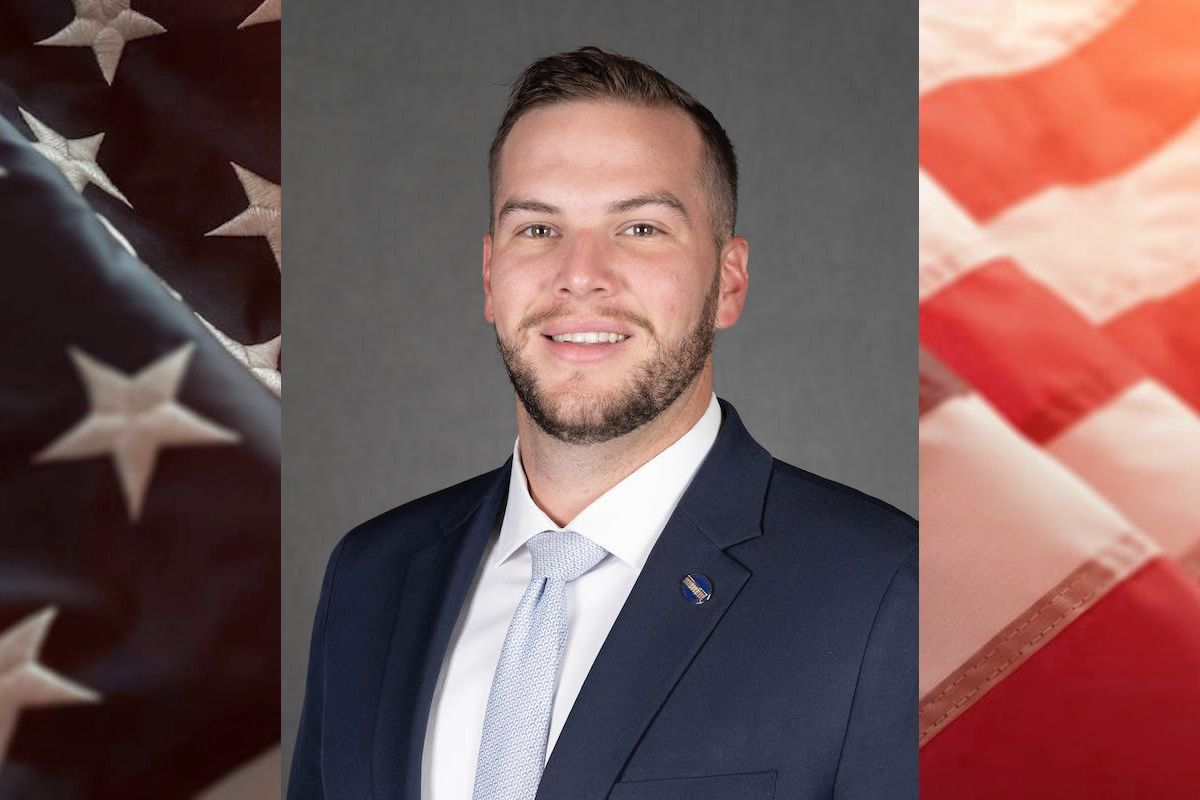 WVU student veteran Ben DeRoos wearing a dark suit and light blue tie. His hair is short and he has a short beard. The photo is laid on top of an American Flag. 