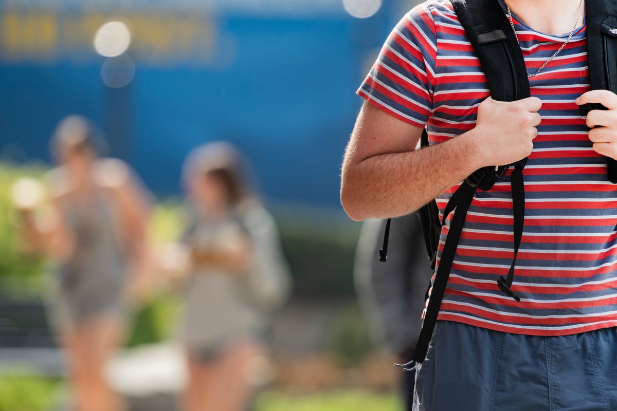 A male student is shown tightly cropped in the foreground. His head is out of the frame but he is wearing a red and blue striped shirt, blue shorts, and a gray and black backpack. His hands are holding the straps on his chest. There are two other people