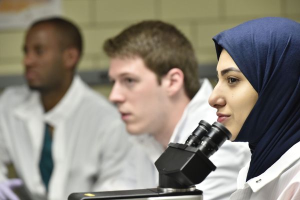 Two male students in white coats blurred in the background and a female student in a navy blue hijab and a white lab coat in focus in front of a microscope.