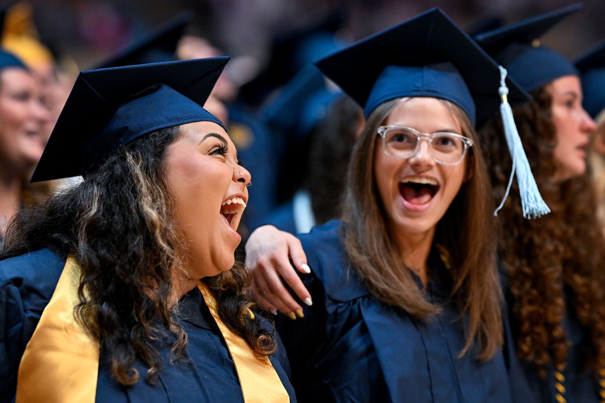 Two graduates wearing navy blue caps and gowns smile with their arms around each other while singing 'Country Roads' to celebrate their graduations.