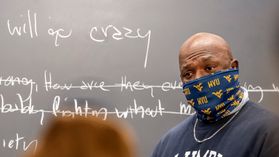 An man in a gold and blue face covering stands in front of a blackboard with chalk writing and some of the writing is crossed out 