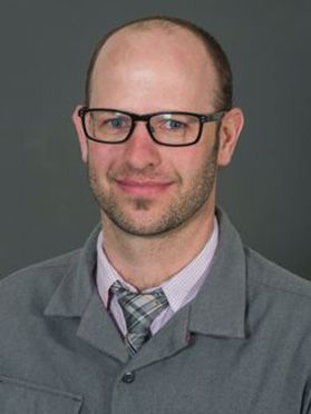 Headshot of WVU professor Nike Zegre. He is pictured against a gray background wearing a gray jacket with a pink dress shirt underneath. He has a receding hairline and is wearing black, square-framed glasses. 