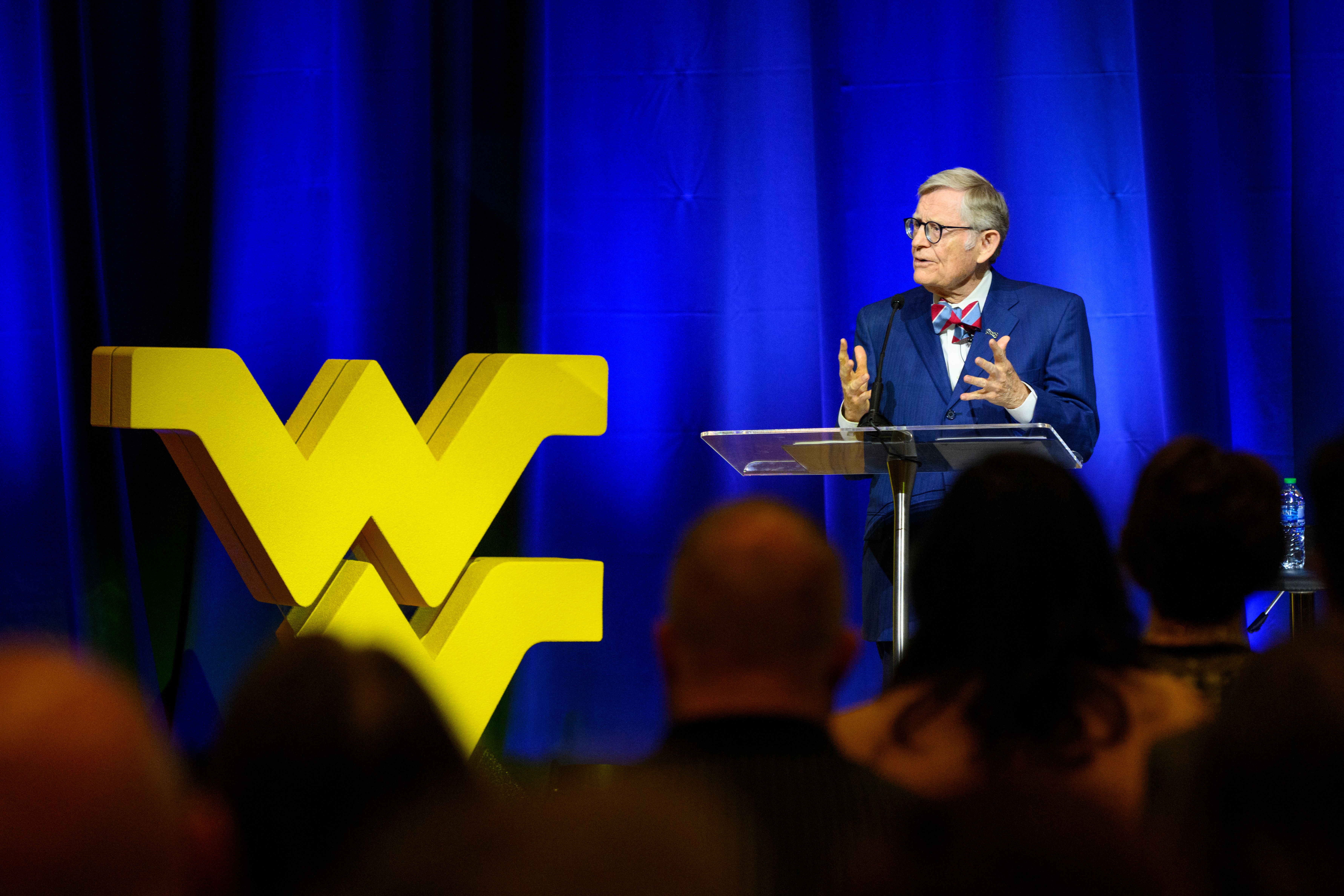 President Gordon Gee stands behind a clear podium in front of a blue curtain. A gold Flying WVU is to his right.