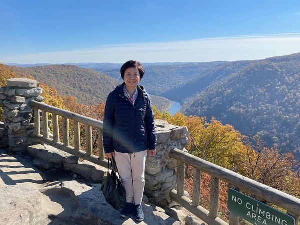 WVU alumna Choalu Chen is pictured here standing over the Cooper's Rock overlook. She is carrying a black purse and is wearing khaki pants with a navy blue coat. She has short, black hair. 