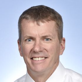 Headshot of WVU researcher Chris Martin. He is pictured against a light blue background and is wearing a white dress shirt. He has short light colored hair. 