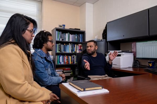 Omar-Abdul Aziz is seated at his desk in his office. He is wearing khaki pants and a navy blue sweater. Two of his students are shown facing him at the desk. Both have dark hair and one is wearing a blue jean jacket. 