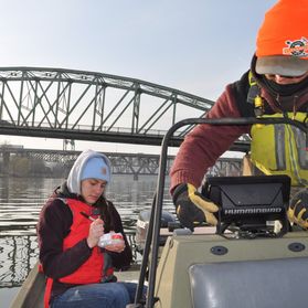 Two WVU researchers bundled up in warm clothing ride in a boat on a river with a span bridge in the background. 