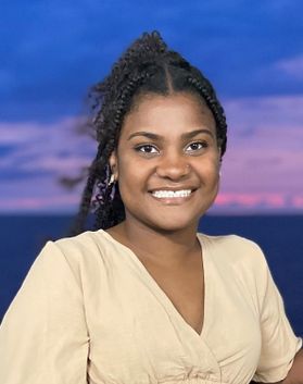 Headshot of WVU student Raiven Scott. She is pictured outside with the ocean behind her. She is wearing a cream colored top and has curly black hair. 