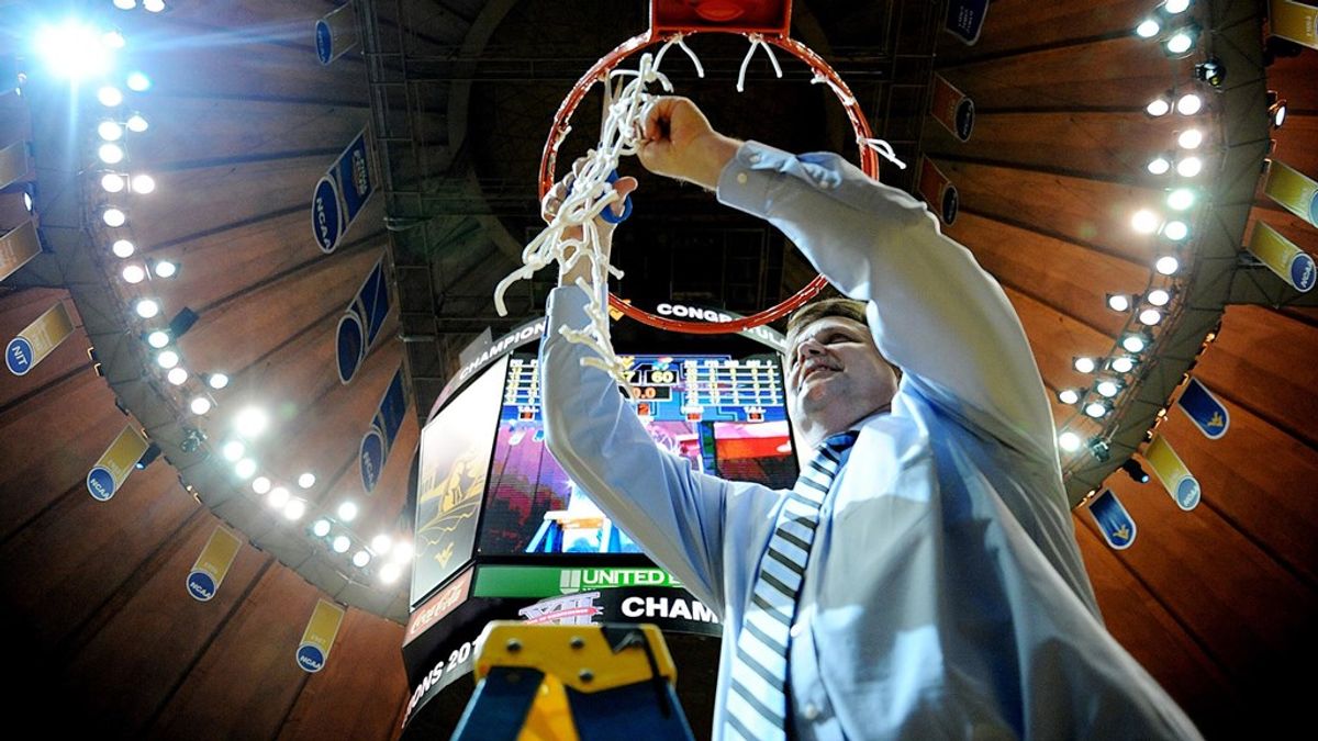 Shot from the floor, this photo shows a man in alight blue shirt and tie cutting down a basketball net under the video board and a circle of lights at WVU's Coliseum