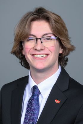 Headshot of WVU Foundation Scholar Colin Street. He is pictured against a gray background wearing a dark colored suit, white dress shirt, and purple patterned necktie. He has shoulder length blonde hair and is wearing square-framed glasses. 