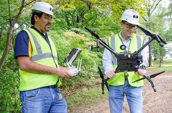 Two people are shown here wearing fluorescent yellow vests and white hard hats. One is holding a controller. The second is holding a drone.