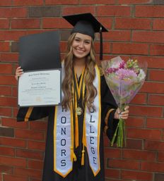 Scholarship recipient Lexi Mullen is pictured wearing a dark colored cap and gown with a white and gold "Honors College" sash. She is holding her diploma and a bouquet of cut flowers.  