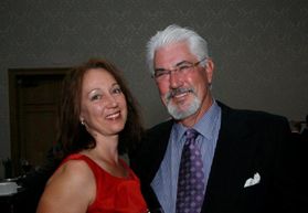 A woman with brown hair wearing a red dress standing next to a man with white hair with glasses wearing a blue suite and tie