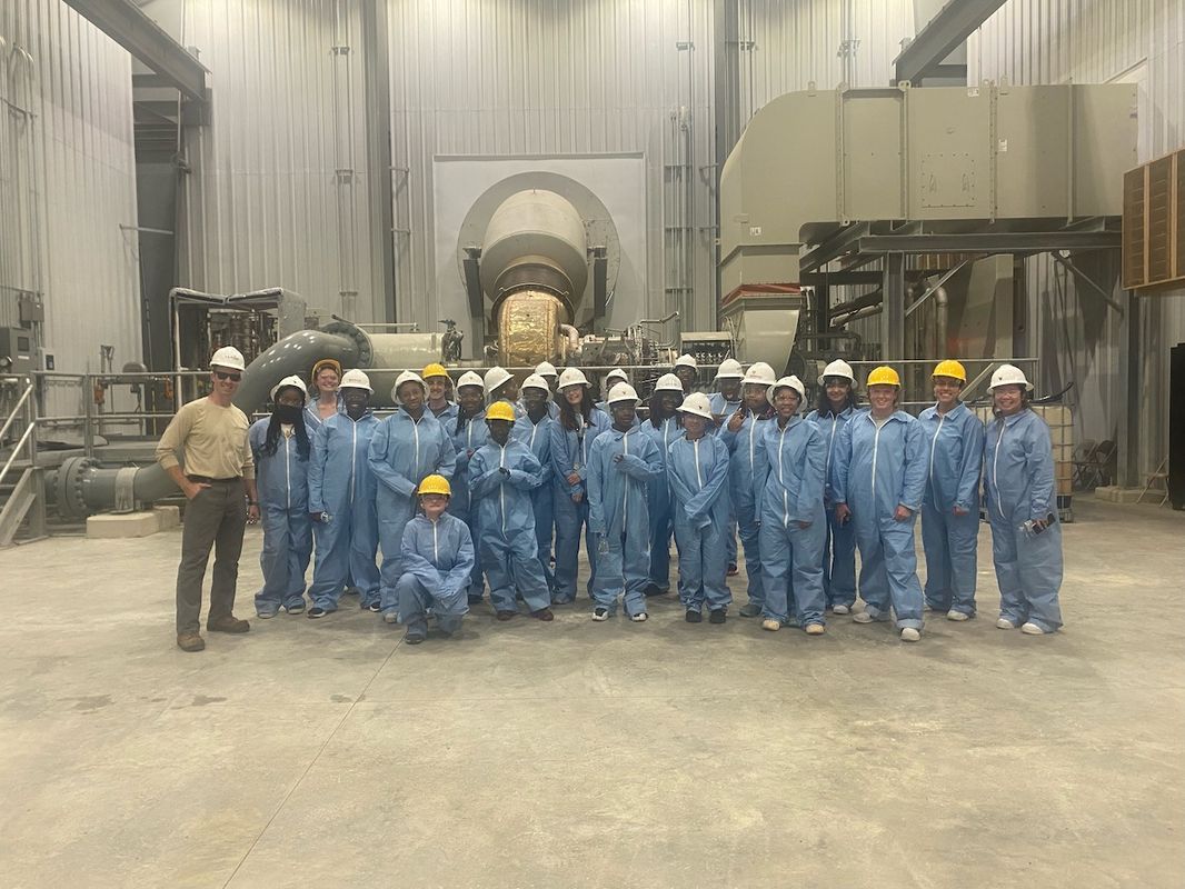 More than a dozen students wearing blue work clothes and hard hats stand in a factory room with gray walls.