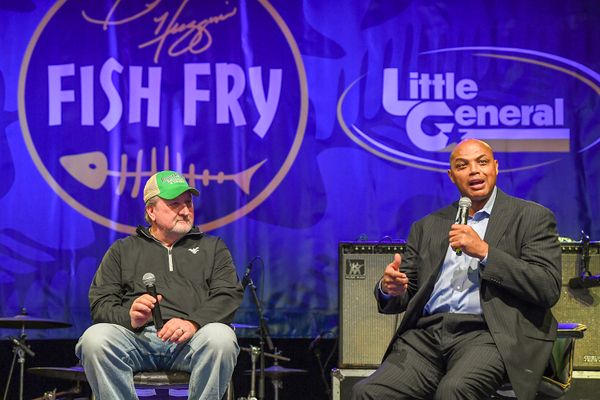 Picture taken at annual Bob Huggins Fishy Fry. In the picture. Huggins, wearing jeans, a gray sweater and a green baseball hat, sits to the left of famous basketball player Charles Barkley who is holding a microphone and talking. He is wearing a gray suit