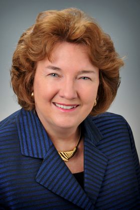 Headshot of outgoing WVU Tech president Carolyn Long. She is pictured against a light gray background. She is wearing a navy blue tonal striped top and a gold necklace. She has shoulder length red hair. 