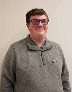Headshot of WVU horticulture student Garrett Oursler. He is pictured standing against a beige wall and is wearing a 3/4 zip sweater and dark framed glasses. He has shortly, light brown hair. 