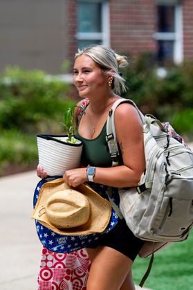 A young female WVU student carrying belongings during move-in week at WVU. She has a gray backpack on her back and is holding a straw cowboy hat and a potted plant. 