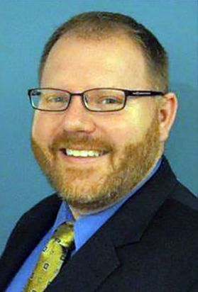 Headshot of WVU administrator Ted Svehlik, He is pictured in front of a light blue background wearing a dark suit jacket over a blue shirt and gold patterned tie. He has light colored hair and a full beard, and wears glasses. 