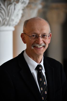 A smiling, partly bald man with glasses wearing a suit coat and shirt and tie