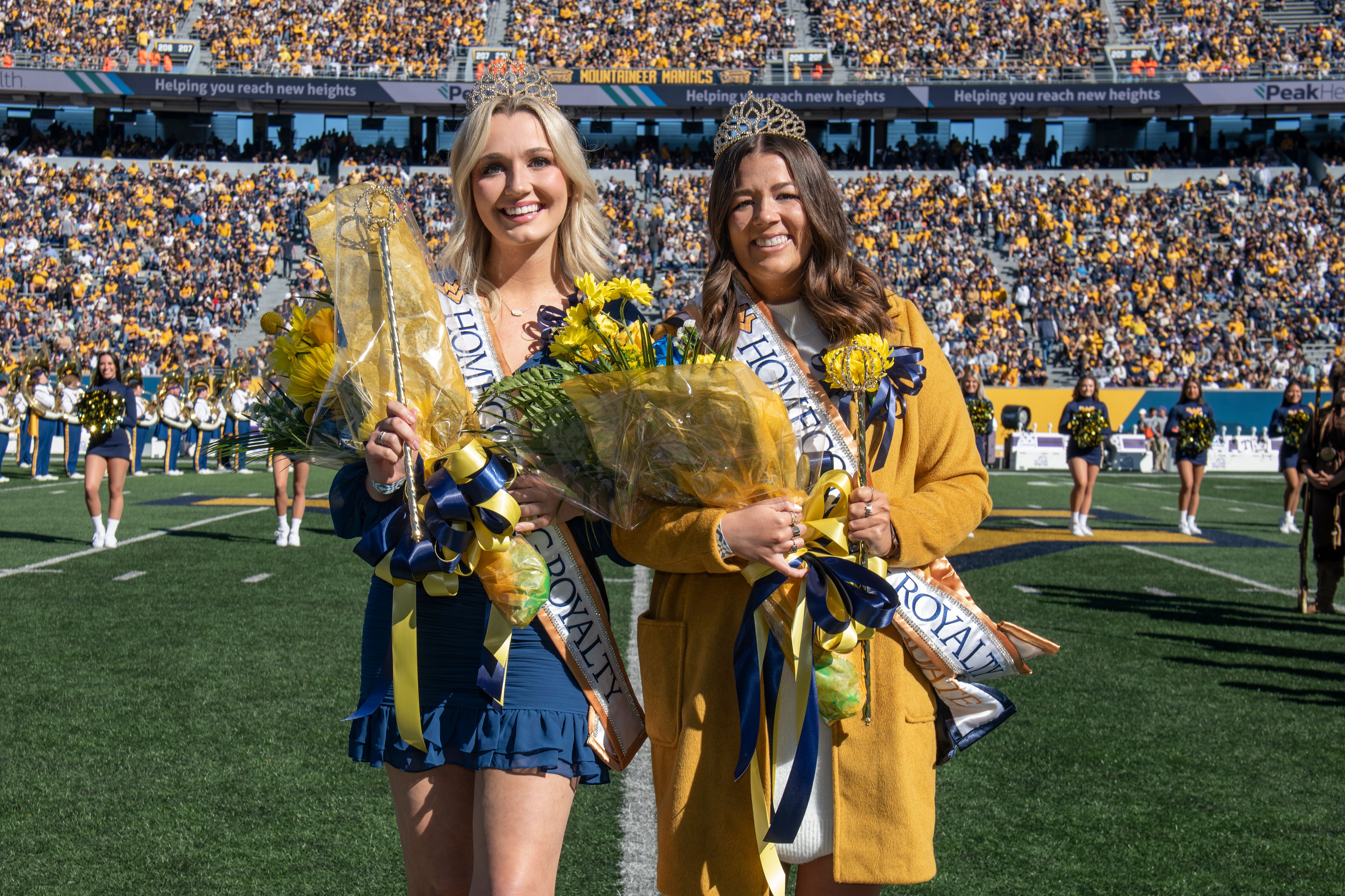 Photo featuring WVU Homecoming Royalty. Two women were chosen for the honor and each are on the football field wearing a crown and holding a large bouquet of flowers. 