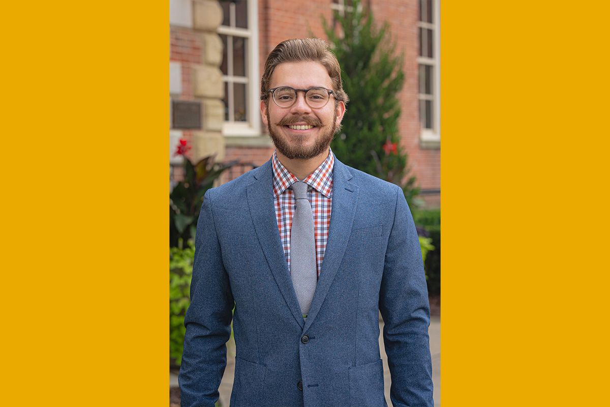 Headshot of WVU graduate student David Malecki. He is dressed in a gray suit and tie standing in Woodburn Circle. He has light brown hair and a full beard. 