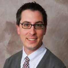 Headshot of WVU researcher Jonathon Beckmeyer. He is pictured indoors and is wearing a white dress shirt, checked tie, and gray v-neck sweater. He had short, styled black hair and is wearing black-rimmed glasses. 