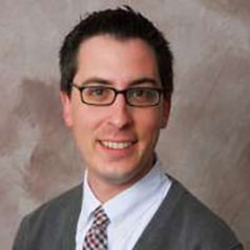 Headshot of Jonathon Beckmeyer. He is pictured in front of a neutral background and is wearing a gray cardigan over a white dress shirt with a checked necktie. He has short dark hair and wears square rimmed glasses. 