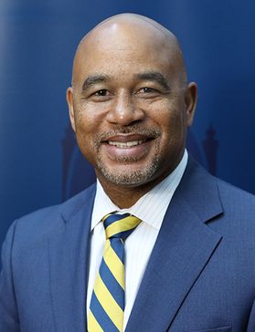 A person stands in front of blue background while wearing a blue jacket and gold and blue tie.