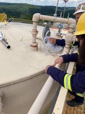 Two researchers wearing safety clothing, hard hats and other safety equipment take samples of vent emissions from a shale gas liquid storage tank. 