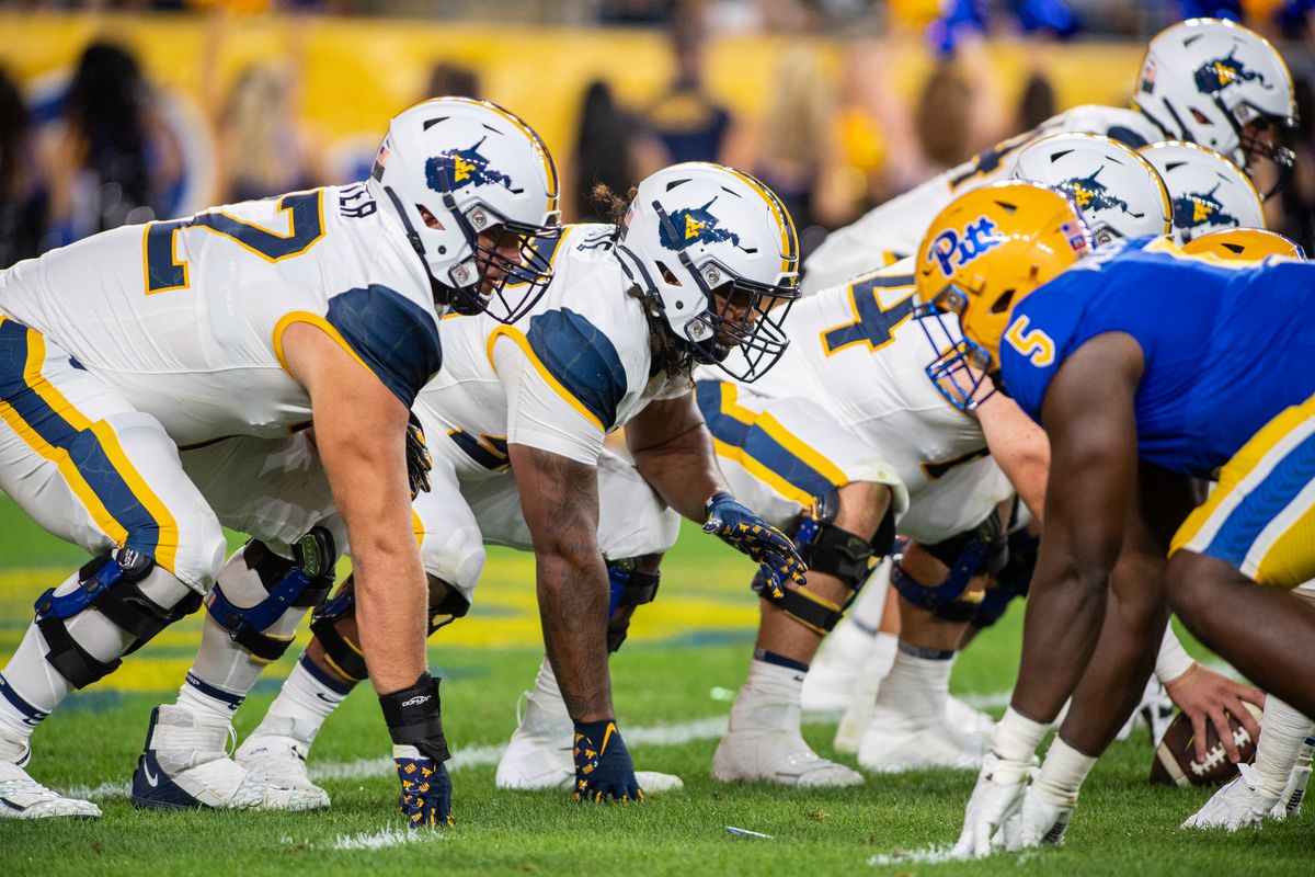 A photograph taken during a WVU football game of players bent down at the line of scrimmage waiting for the ball to be snapped. The WVU players are dressed in white uniforms and the opposing team is Pitt. 