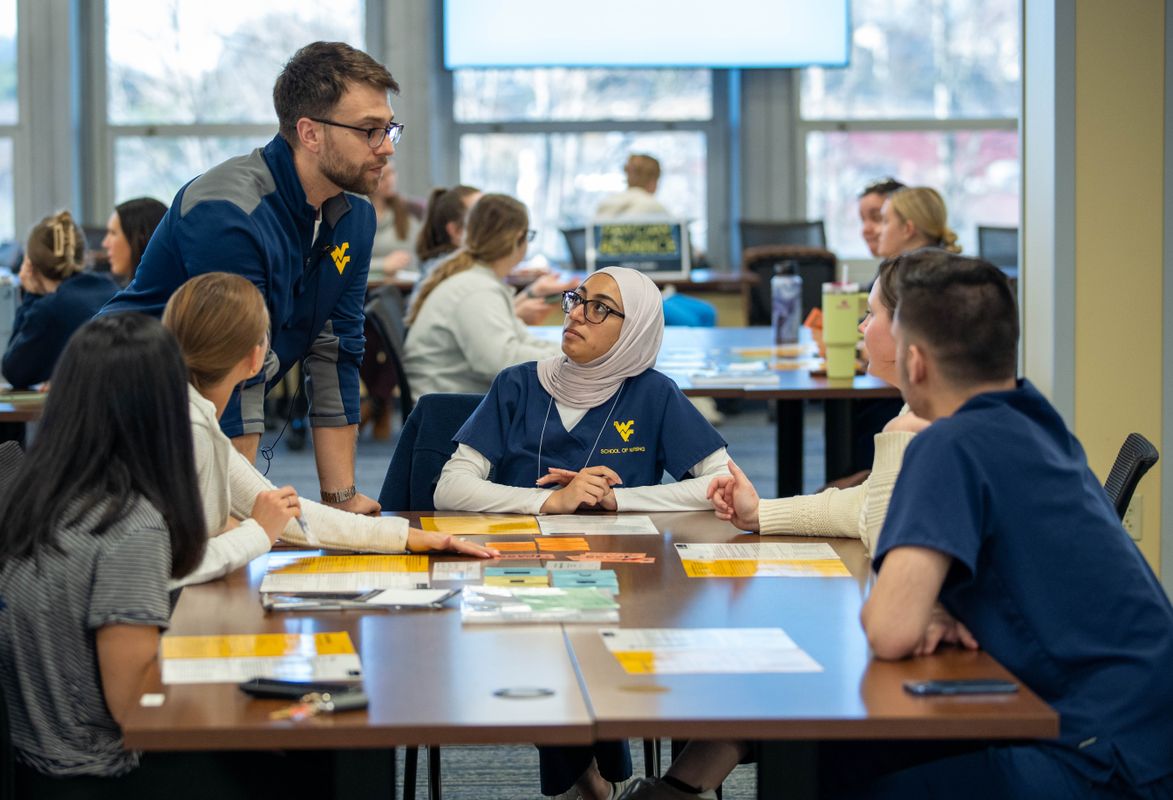 Four students sit around a square wooden table looking at a person standing in the left corner of the table as part of a povery simulation at WVU Health Sciences.