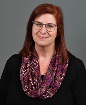 A headshot of WVU employee Lori Hostuttler. She is photographed in front of a gray background wearing a black shirt with a purple patterned scarf. She has shoulder length red hair and wears glasses. 