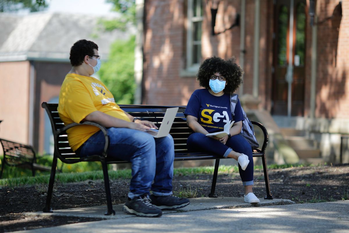 two students sit on a bench outside with masks on
