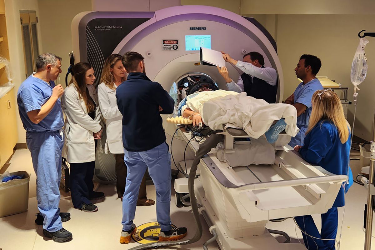 A patient is shown on a hospital bed in front of a larger machine with seven medical professionals watching during a new kind of Alzheimer's treatment.