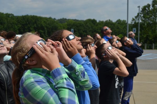 Young students stand in a line while holding glasses to their faces, looking up at the sky.